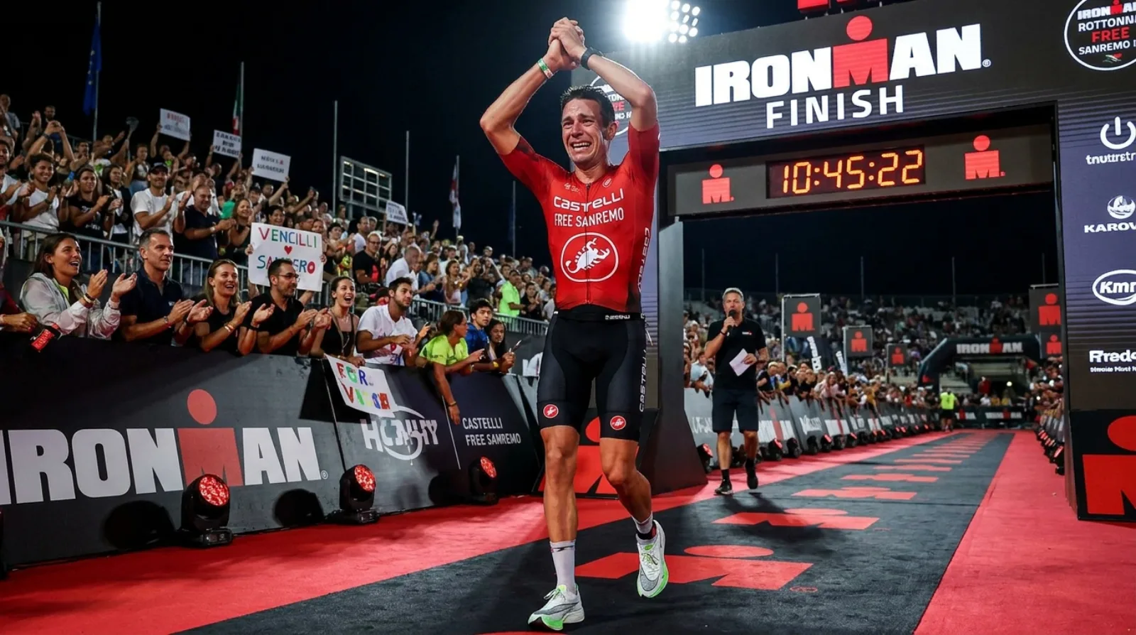 Triathlete crossing the IRONMAN finish line with arms raised in triumph, emotional finish line moment