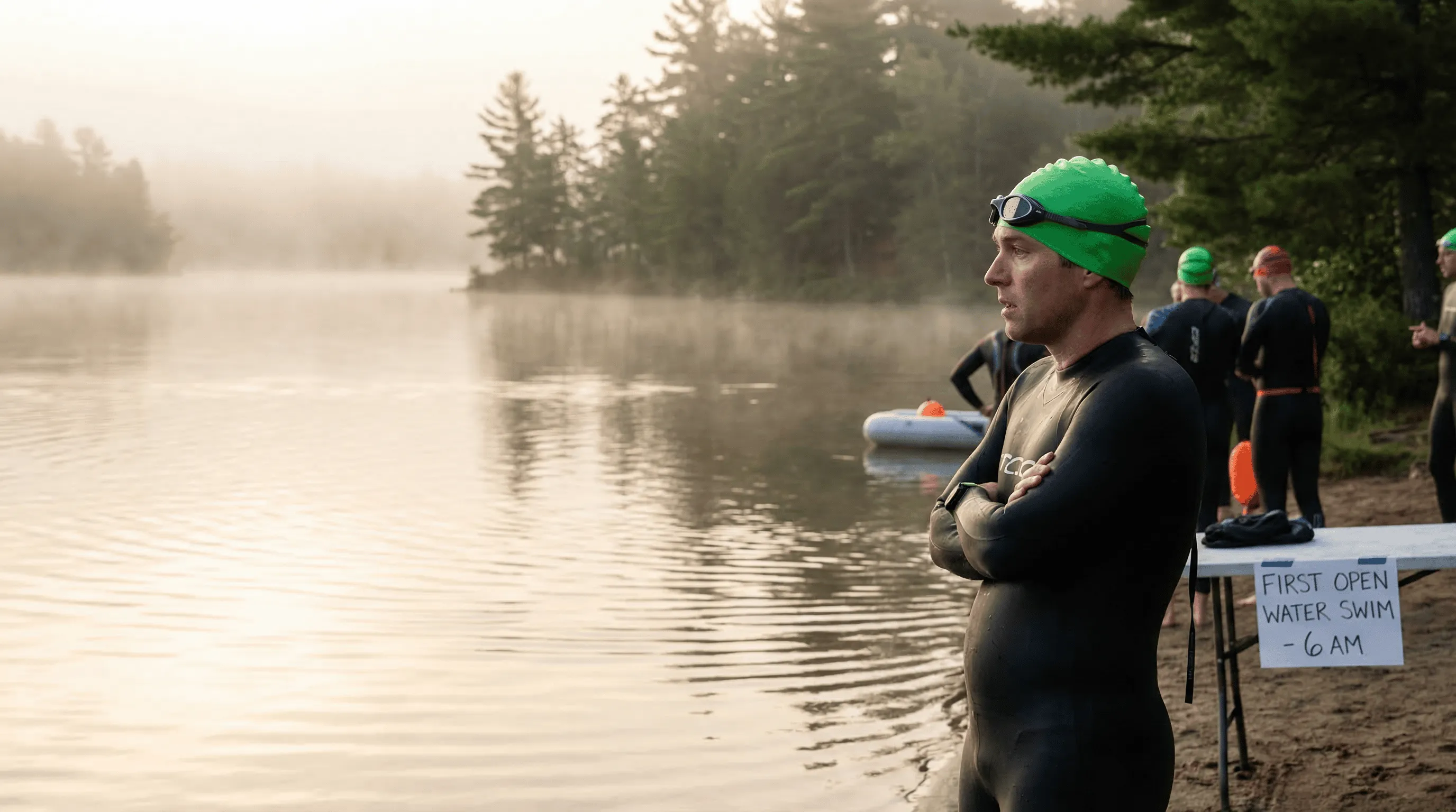 Beginner triathlete in wetsuit standing at the edge of a natural lake at dawn, misty morning light reflecting on the still water