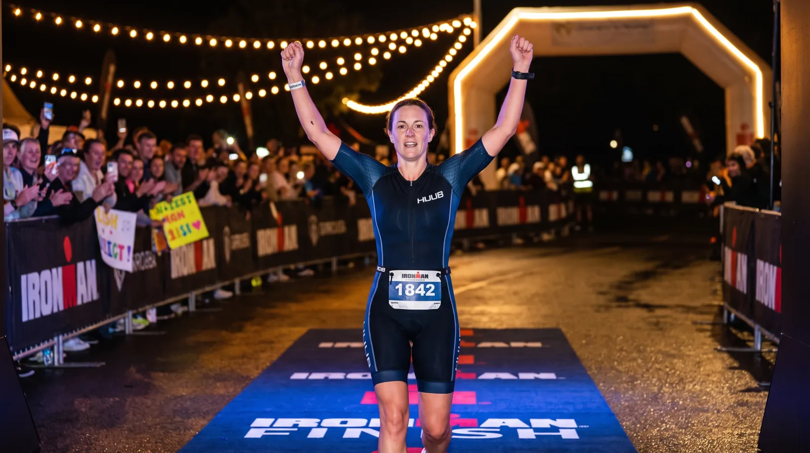 A triathlete crossing the finish line of a full-distance IRONMAN race at night with arms raised and finisher medal lights in the background