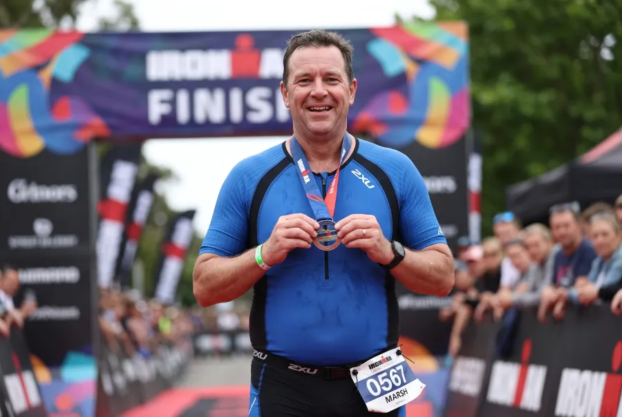 A middle-aged average-looking triathlete smiling and celebrating after finishing their first full-distance IRONMAN race, finisher medal around their neck