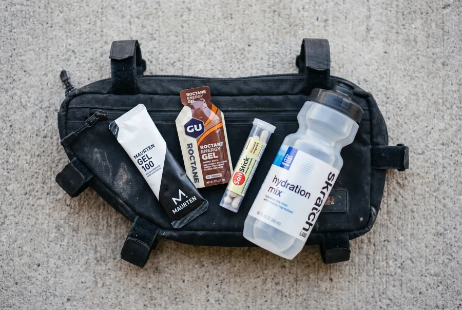 A flat lay of IRONMAN race nutrition products including energy gels, electrolyte chews, and a sports drink mix bottle with visible real brand labels, arranged on a bike top tube bag