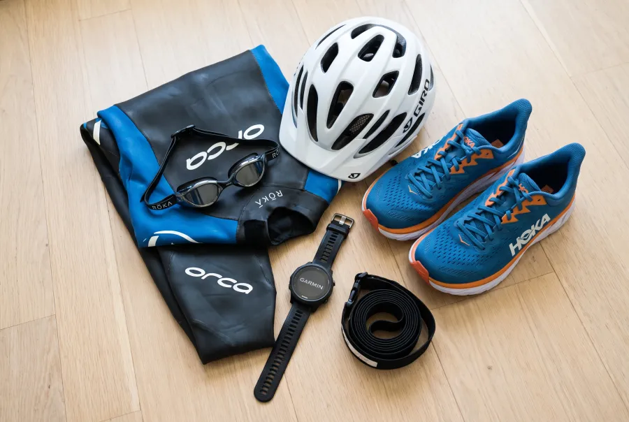 A flat lay of first-time IRONMAN gear including a wetsuit, swim goggles, a cycling helmet, running shoes, and a GPS triathlon watch, with visible real brand logos on each item