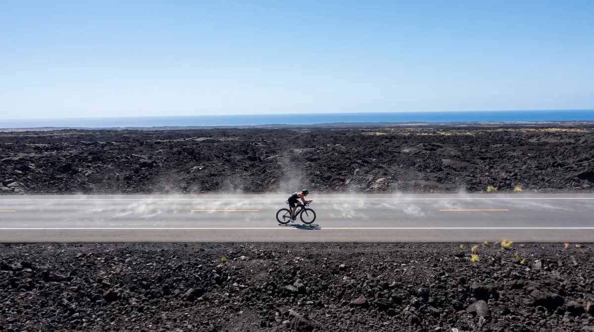 A lone cyclist riding across the black lava fields of the Queen Kaahumanu Highway near Kona, Hawaii, with the ocean visible in the distance