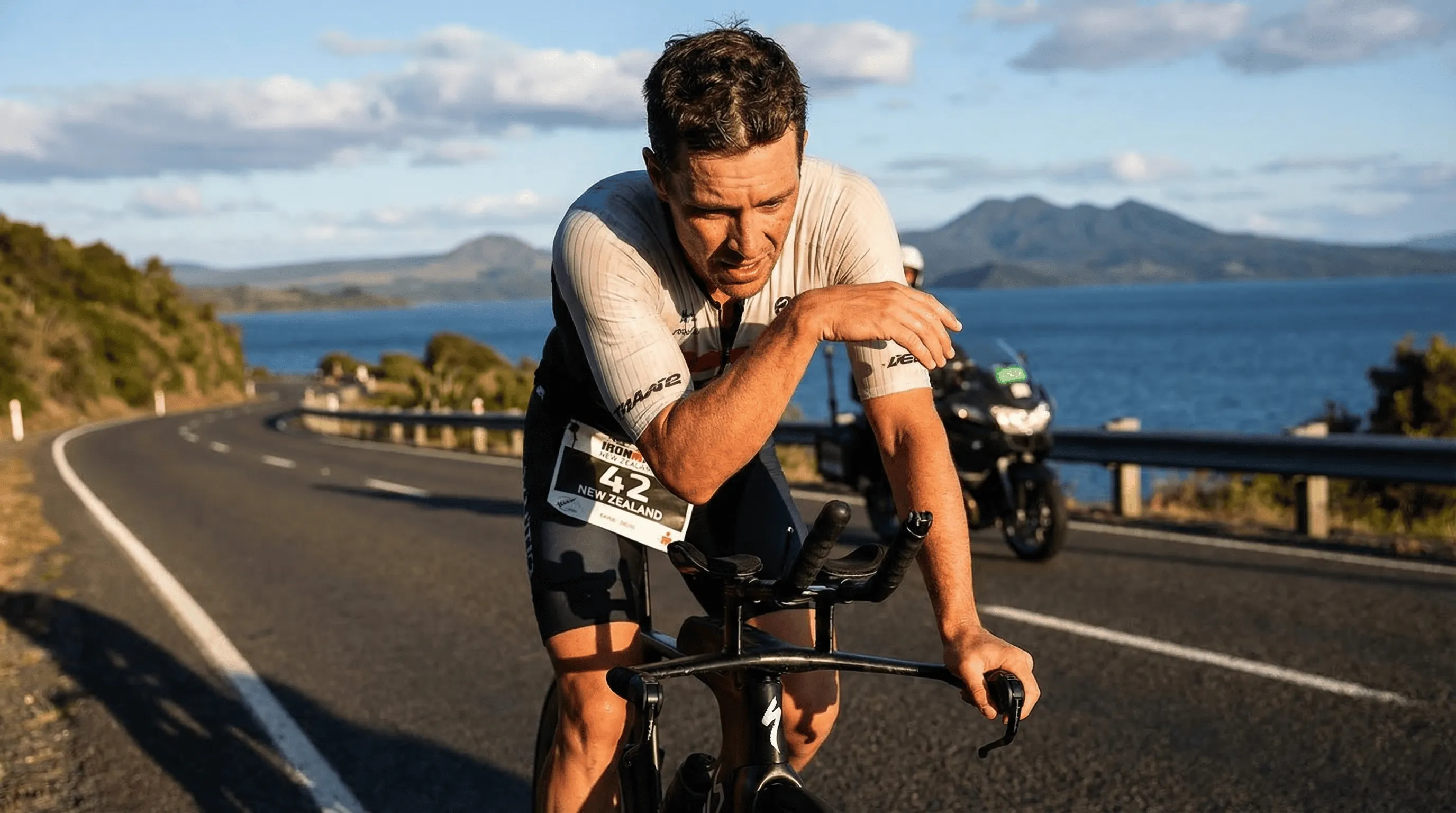 Professional triathlete riding a TT bike on a scenic New Zealand coastal highway with Lake Taupo in background