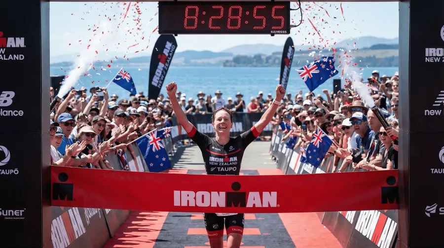Professional triathlete crossing the Ironman New Zealand finish line in triumph with crowd cheering and timing clock showing 8:28:55