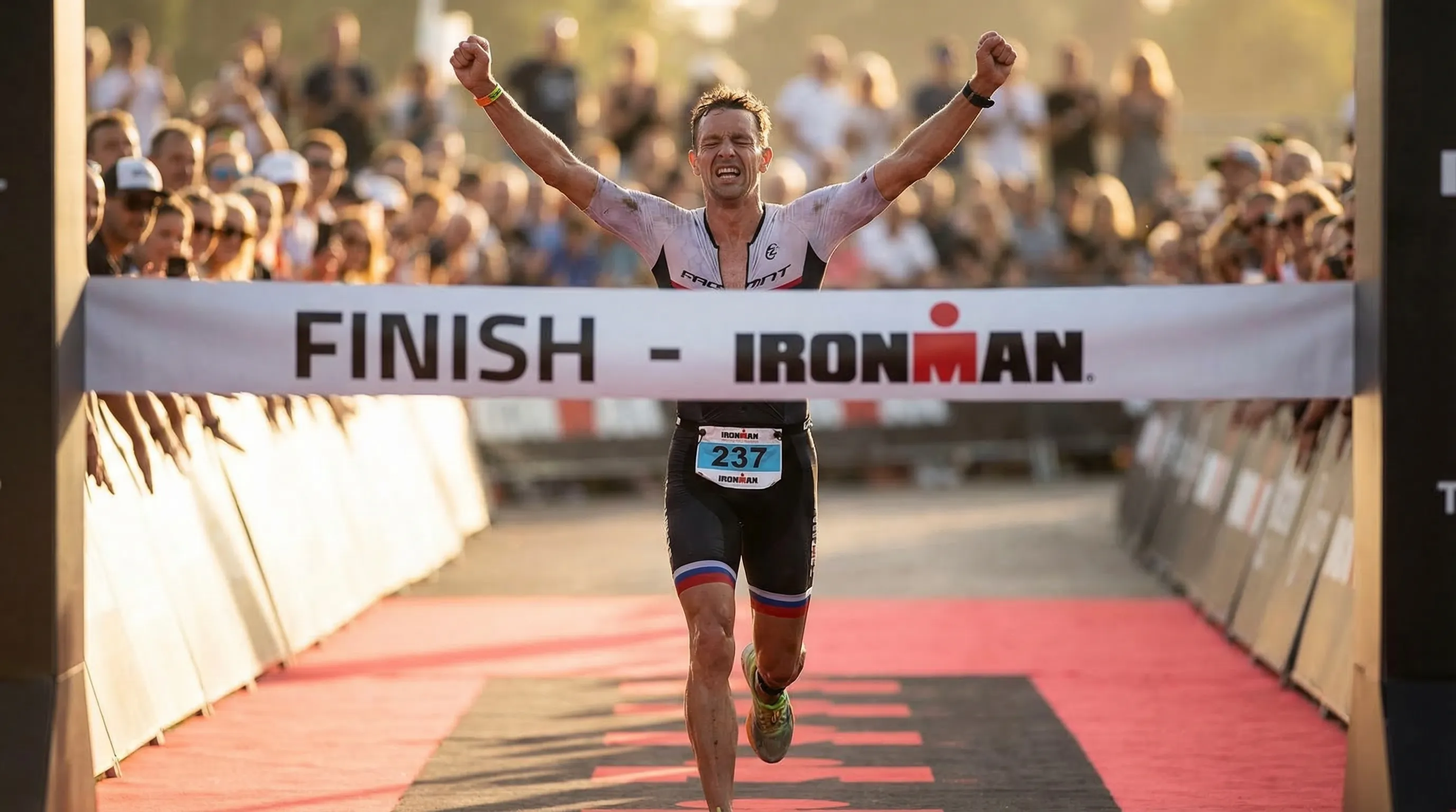 Triathlete crossing a finish line with arms raised in triumph, exhausted but elated, cheering crowd behind
